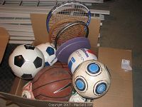 Collection of assorted sports balls and badminton racquets in a cardboard box, showing a variety of colors and types including soccer balls, basketball, and shuttlecock.