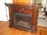 Frontal view of electric fireplace with clear glass front, faux logs inside, and dark wood mantel with decorative carved columns and floral medallion.