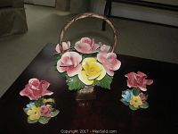 Three Capodimonte floral porcelain pieces on a dark wooden table; one large basket centerpiece and two smaller floral figurines