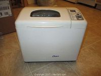 White Oster bread machine, model 5834, shown from the front side with brand logo visible, placed on tile floor.