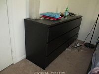 Front-facing photo of the black IKEA dresser showing six drawers with no handles, placed against a white wall with various items on top and connecting cables on the floor.