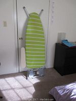 Full length view of the ironing board standing upright showing the striped cover and attached sleeve board on the left side.