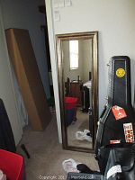 Full length mirror 17x53, with gold tone frame leaning against a wall next to black instrument cases and a red container.