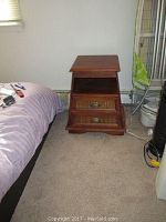 Side view of wooden side table placed next to bed, showing two cane front drawers with brass handles and an open shelf above.