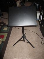 Full view of the black music stand with tripod legs and rectangular sheet holder on carpeted floor near furniture.