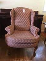 Front view of the wing chair showing the pink diamond patterned upholstery, cushioned seat, rolled arms, and wooden cabriole legs.