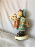 Side profile view of the Hummel figurine showing the mail boy holding a love letter with a red heart seal, wearing blue coat and cap with brown shoes.