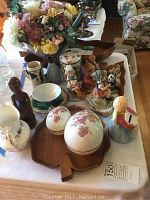 Photo showing a table with an artificial flower arrangement, wooden leaf bowls, floral porcelain covered dishes, ceramic pitcher, wooden figurine carving, and several small figurines grouped.
