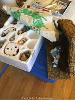 Nativity figurines in labeled white foam storage container with paper cover and a moss-textured stable beside it