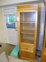 Full view of tall wooden cabinet with glass door, showing door handle, shelves inside, and bottom drawer. Positioned near a door and some furniture.