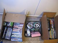 Three cardboard boxes filled with assorted CDs, DVDs, VHS tapes, and video game cases.