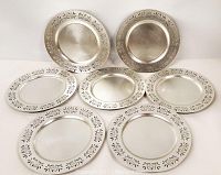Full view of seven silver-tone charger plates arranged in a group, showing front surfaces and decorative etched floral pattern around edges.