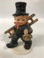 Front view of the Hummel chimney sweep figurine showing facial features, black hat, black outfit, red scarf and ladder across shoulder.
