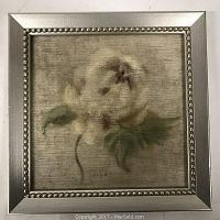 Framed print showing the full artwork of a pale flower with green leaf on a neutral background, in a metallic silver frame with beaded edge.