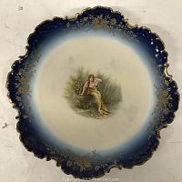 Top-down view of 9-inch Bavaria flow blue porcelain plate showing central image of seated young woman and graduated blue and gold decorative rim.