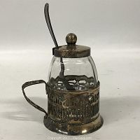 Front angled view of a small glass mustard pot in a silver plated holder with handle and matching hinged lid and spoon.