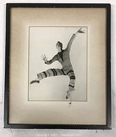 Framed black and white photograph showing a ballet dancer in mid-air leap wearing a striped costume. The frame is black with an off-white mat.