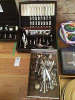 Full view of various pieces of silver-tone flatware including spoons, knives, forks, serving utensils, and napkin rings displayed on a table with some in boxes and some loose.