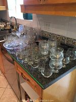 Wide view showing multiple glassware items arranged on a granite countertop, including wine glasses, shot glasses, pitchers, and serving tray.
