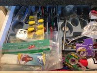Overview photo showing various kitchen and household items including gray cutlery trays with small hand tools such as screwdrivers, yellow-handled kitchen items, and plastic food storage bags and wraps spread across the trays.