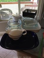 Photo showing various clear Pyrex glass baking dishes including rectangular and square pans, a white covered ceramic bowl, black rectangular metal pan, and a white ceramic gravy boat.