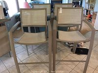 Front view of two metal frame bar stools showing woven beige seats and backs with armrests.