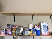 Wide shot of shelf with assorted household supplies including garbage bags, aluminum roasting pans, aluminum foil, Lysol wipes, disposable napkins, and a blue plastic insulated cooler jug.