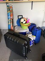 Photo showing assembled camping gear including Samsonite suitcase in front, Coleman cooler behind it, two blue sleeping bags on the right, a propane tank, a green plastic pail with toys, and two umbrellas standing upright in the back