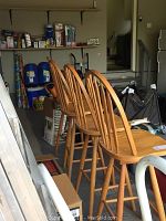 Four wooden bar stools with vertical slatted backs lined up in a cluttered garage space, showing overall design and condition.