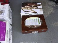 Closed wood-finished carrying case with metal handle and key lock, packaging box behind it.