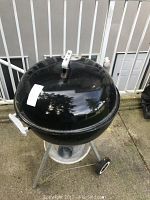 Full view of black Weber dome charcoal kettle grill on tripod stand with two wheels, showing overall size and conditioning outdoors on concrete.