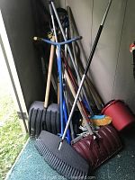 Assorted garden tools leaning against wall inside storage area including multiple snow shovels, rakes, broom, and bucket