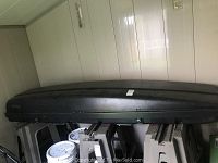 Side view of the black Thule Summit car top carrier showing overall shape and size. Positioned horizontally on plastic step stools with parts of wall and paint bucket visible.