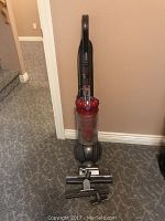 Full view of upright vacuum standing on carpet showing body, handle, dust bin, hose coiled on side and base ball wheel