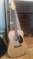 Full front view of the Jasmine acoustic guitar standing next to a cardboard box and furniture, showing the natural wood finish and black pickguard.