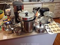 Wide view showing coffee maker, pots, pans, kettle, toaster, flour sifter, muffin tin, glass bowl, and canned seasoning on kitchen counter.