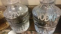 Photo showing two clear cut glass round liquor decanters with metal labels GIN and VODKA