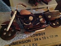 Close-up image of largest wooden motorcycle model in dark and light polished wood. Features detailed wheels, handlebars, and engine parts. Box beneath labeled 'LOWBOY'.