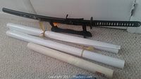 Sword resting on black decorative stand with rolled maps below it to the left on beige carpet.