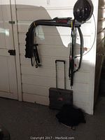 Folding bike rack with helmet, rolling suitcase, and protective guard hanging on wall.