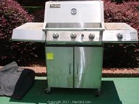 Full front view of Charglow gas grill with four burner knobs, two door cabinet below, stainless steel side panels, and black grill cover on the ground beside it.