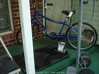 Blue Fuji Saratoga 3.0 men's bicycle on porch, showing frame, seat, handlebars, and rear tire, with black floor mats underneath.