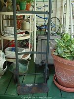 Black metal hand truck with two wheels and curved handle positioned near other lot items on porch deck, including a large clay pot with live plants.