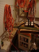 Photo showing multiple long orange extension cords hanging on pegboard, some coiled and some loosely hanging, with part of a Juiceman juicer box visible beneath.