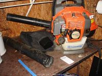 Husqvarna 130BT gas leaf blower with two detached black tubes and a black fabric collection bag on a table.
