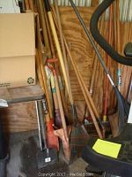 Photo showing garden tools including shovel, rakes, and poles leaning against wall next to metal sawhorse with wooden top, some cardboard boxes visible