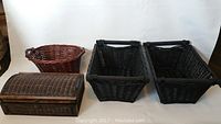 Photo showing two rectangular black wicker baskets and one small brown wicker chest with hinged lid, all placed on a white surface against a white background.