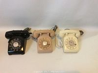 Front view of three rotary phones in black, cream, and pink colors displayed side by side, showing full phones with rotary dials and handset cords coiled.
