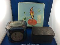 Three vintage metal tins placed together on a blue background. The largest tin is square with a bird image, the second is a blue octagonal tin with a floral and bird design, and the third is smaller, rectangular and embossed.