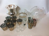 Photo showing entire glassware set including silver wine cooler, multiple shot glasses, fish-shaped glass, tall glasses with geometric designs, and clear gold-rimmed tumbler.